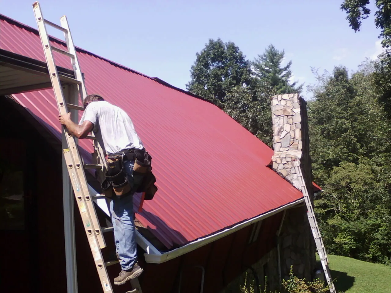 Roofer on a ladder beside a red metal roof and stone chimney — Soffit & Fascia Repair by Roofing Brothers in Ogunquit